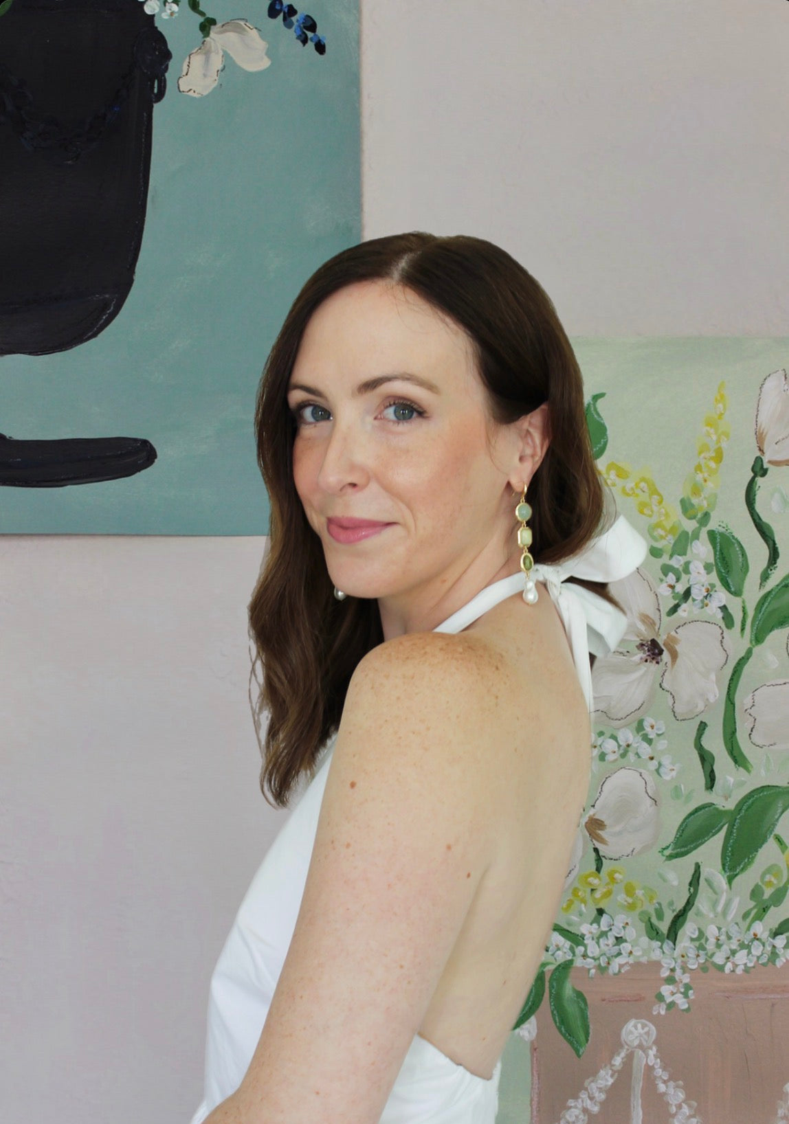 A woman with brown hair wearing a white halter dress with a bow at the back, posing in front of a floral painting.